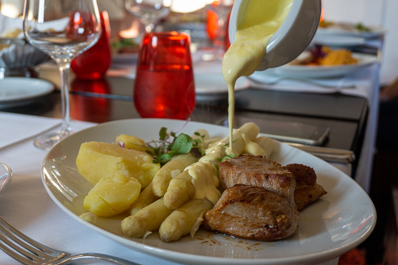 Auf einem ovalen Teller ist ein Menü mit Spargel, Kartoffeln und Fleisch angerichtet. Aus einer Sauciere wird Sauce Hollandaise ausgegossen.