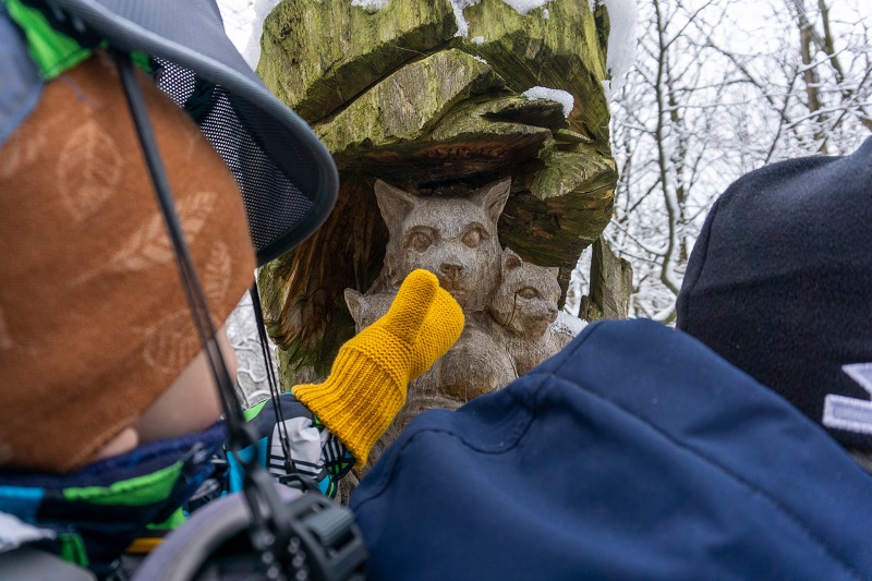 Ein kleines Kind in einer Trage greift nach dem Gesicht einer aus Holz geschnitzten Wildkatze. Das Kind trägt dabei gelbe Woll-Handschuhe.