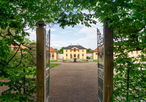 Blick durch eine Hecke mit geöffnetem eisernen Tor auf das Hauptgebäude der Orangerie des Schlosspark Belvedere.