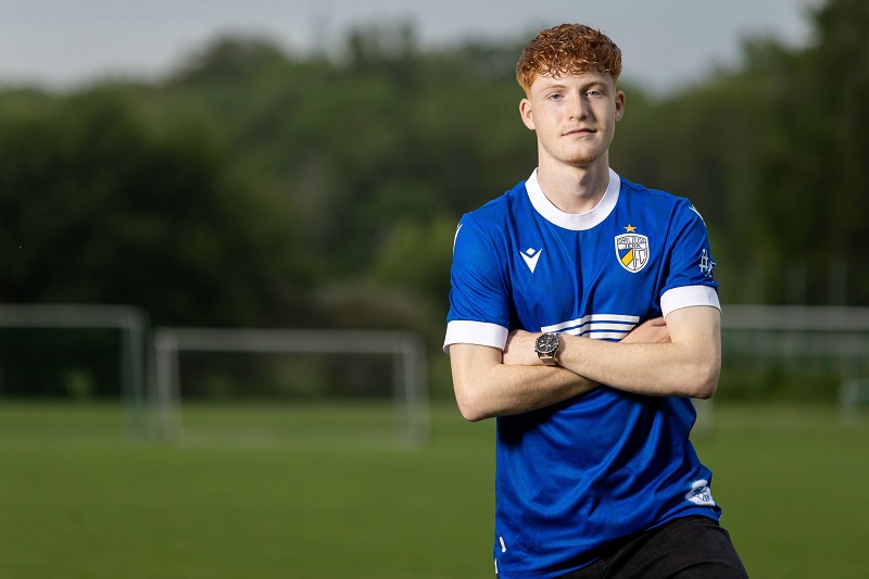 Portrait von Justin Smyla, der mit verschränkten Armen und im blauen Fußballtrikot auf dem Sportplatz steht.