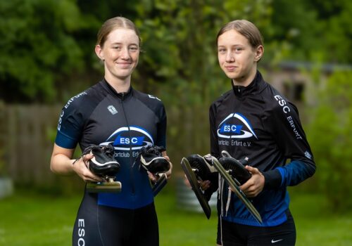 Die Schwestern Paula und Carla Albrecht stehen in ihrer Trainingskleidung nebeneinander und halten beide ihre Schlittschuhe in der Hand.