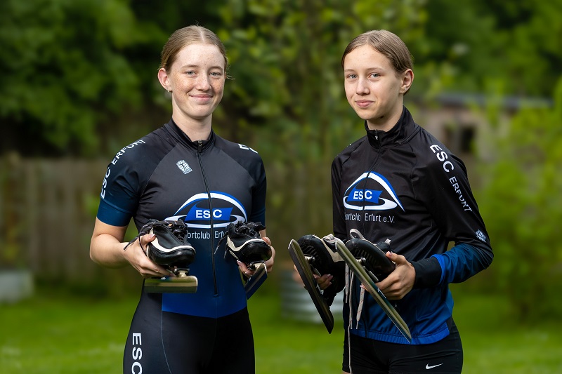 Die Schwestern Paula und Carla Albrecht stehen in ihrer Trainingskleidung nebeneinander und halten beide ihre Schlittschuhe in der Hand.