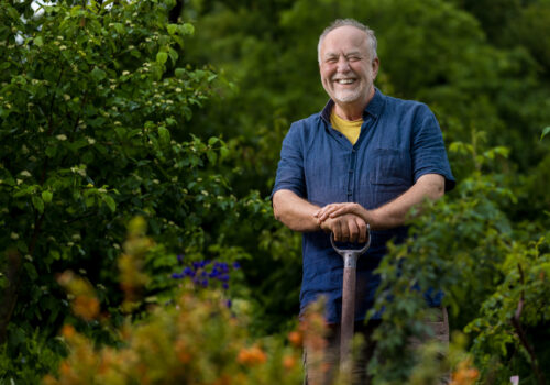 Portrait von Michael Dane. Er steht auf einer Schaufel gelehnt in seinem Garten in Tiefengruben zwischen Sträuchern und Büschen.