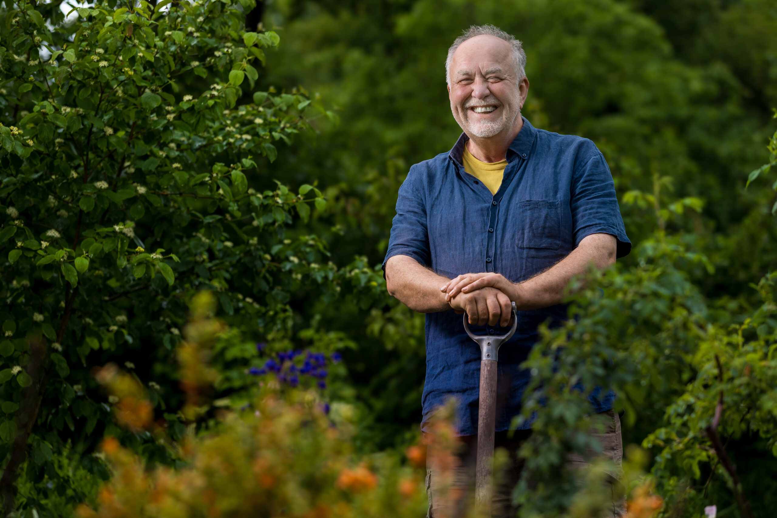 Portrait von Michael Dane. Er steht auf einer Schaufel gelehnt in seinem Garten in Tiefengruben zwischen Sträuchern und Büschen.